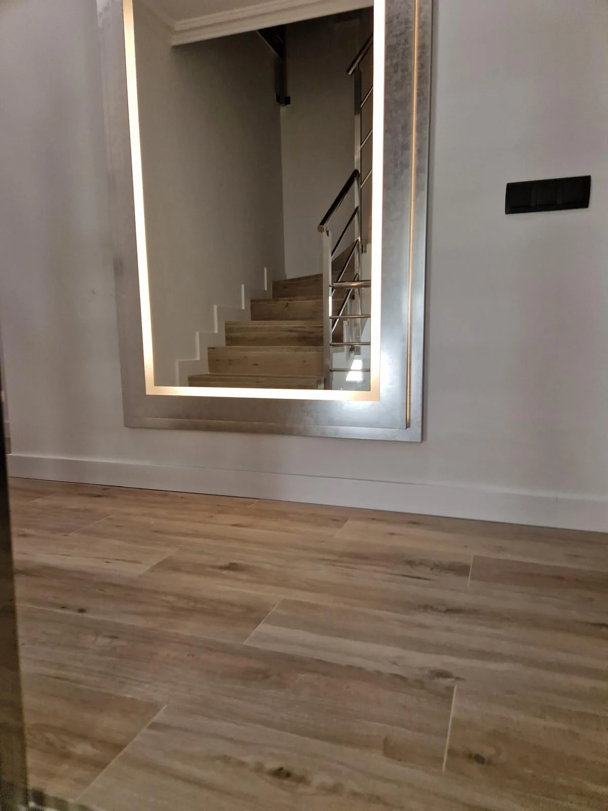 Espejo alto con marco plateado y luz LED integrada, junto a la escalera de una casa