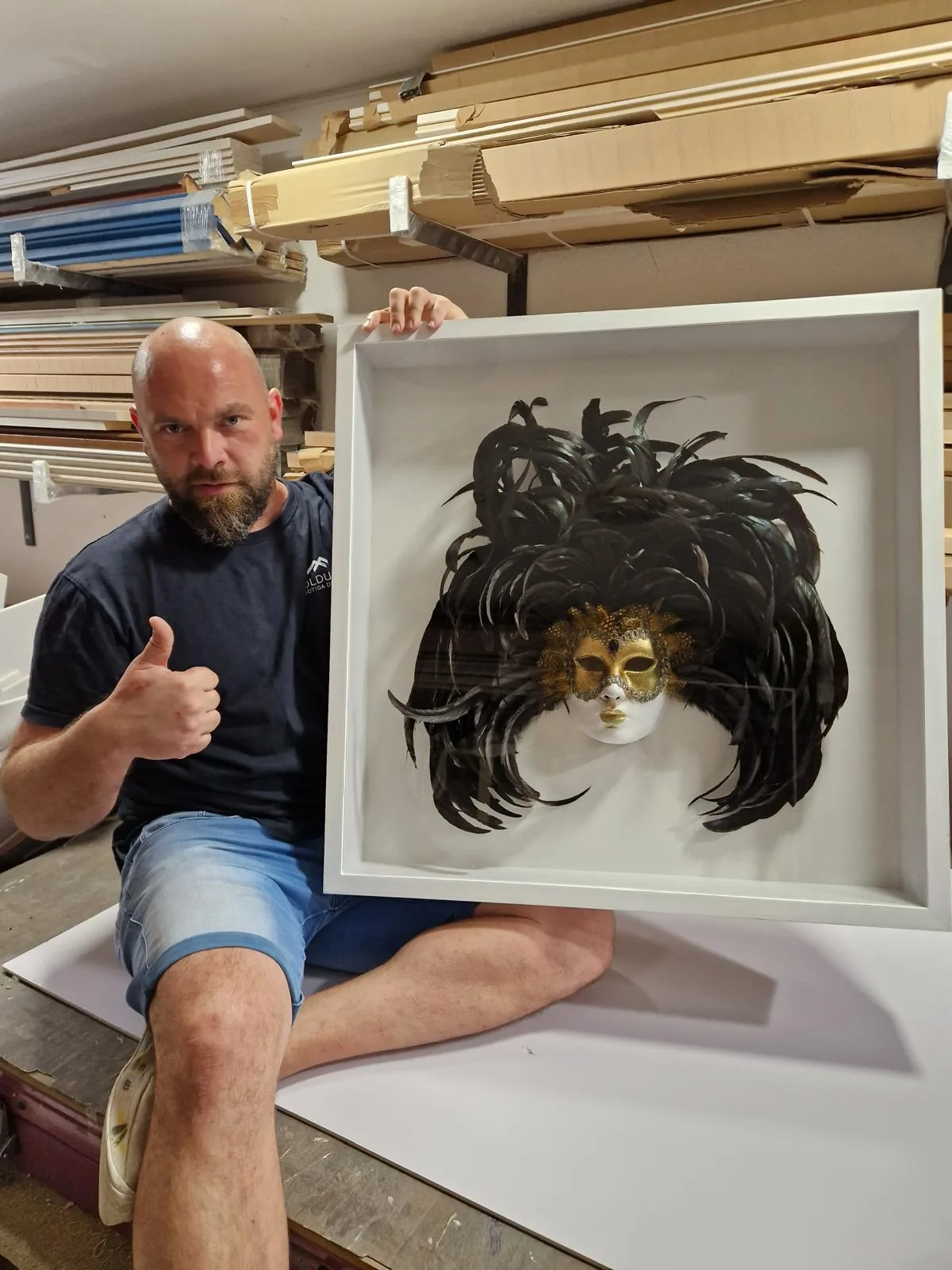Máscara veneciana de plumas negras montada en una vitrina blanca de fondo profundo, en el taller